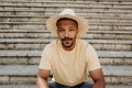 Stylish dark-skinned confident man in trendy straw hat sitting on the stairs. African hipster treveler relaxing at city Royalty Free Stock Photo