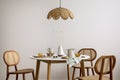 The stylish dining room with round table, rattan chair, lamp and kitchen accessories. Green leaf in vase. Beige wall. Home decor