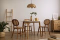 The stylish dining room with round table, rattan chair, wooden commode, pock up poster and kitchen accessories. Beige wall with