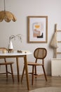 The stylish dining room with round table, rattan chair, wooden commode, pock up poster and kitchen accessories. Beige wall with