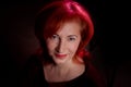 Stylish elderly woman with red hair in vinous dress posing in studio with dark background and red flash and light