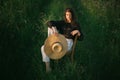 Stylish elegant girl holding straw hat and sitting on rustic chair in summer green field in evening sunlight. Fashionable young Royalty Free Stock Photo