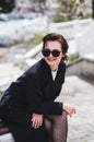 Stylish elegant happy woman sitting on the bench. She dressed in dark coat and sunglasses Royalty Free Stock Photo