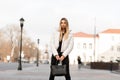 Stylish fashion model of a young woman in a white fashionable jacket in stylish jeans with a leather trendy bag stands on the Royalty Free Stock Photo