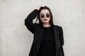 Stylish fine young woman with sexy lips in fashionable round sunglasses in black vintage blazer in trendy t-shirt stands near