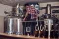 Stylish full bearded Indian man in a fleece shirt and apron controls the brewing process, standing near beer tank in the Royalty Free Stock Photo