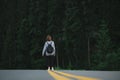 Stylish girl in casual clothes and a backpack walking on the asphalted forest road with yellow markings. Hiker woman walking on a Royalty Free Stock Photo