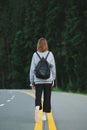 Stylish girl in casual clothes and a backpack walking on the asphalted forest road with yellow markings, vertical photo. Hiker