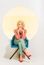 Stylish girl in colorful clothes looking at camera and sitting in armchair on white with yellow circle