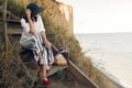 Stylish girl in fashionable look sitting on wooden stairs on beach cliff at sea in sunny light. Happy young boho woman in hat with Royalty Free Stock Photo