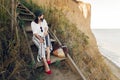 Stylish girl in fashionable look sitting on wooden stairs on beach cliff at sea in sunny light. Happy young boho woman in hat with Royalty Free Stock Photo