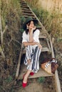 Stylish girl in fashionable look sitting on wooden stairs on beach cliff at sea in sunny light. Happy young boho woman in hat with Royalty Free Stock Photo