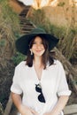 Stylish girl in fashionable look sitting on wooden stairs on beach cliff at sea in sunny light. Happy young boho woman in hat