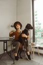 Stylish girl in a hat and with curly hair holds a cup of coffee and hugs the dog in a cozy light cafe, poses for the camera. Lady