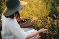 Stylish girl in linen dress gathering flowers in rustic straw basket, sitting in poppy meadow in sunset. Boho woman in hat Royalty Free Stock Photo