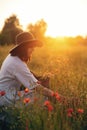 Stylish girl in linen dress gathering flowers in rustic straw basket, sitting in poppy meadow in sunset. Boho woman in hat Royalty Free Stock Photo