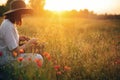 Stylish girl in linen dress gathering flowers in rustic straw basket, sitting in poppy meadow in sunset. Boho woman in hat Royalty Free Stock Photo