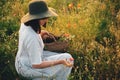 Stylish girl in linen dress gathering flowers in rustic straw basket, sitting in poppy meadow in sunset. Boho woman in hat Royalty Free Stock Photo