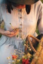 Stylish girl in linen dress gathering flowers in rustic straw basket, sitting in poppy meadow in sunset. Boho woman holding Royalty Free Stock Photo