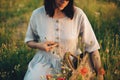 Stylish girl in linen dress gathering flowers in rustic straw basket, sitting in poppy meadow in sunset. Boho woman holding Royalty Free Stock Photo
