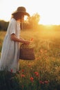 Stylish girl in linen dress gathering flowers in rustic straw basket, walking in poppy meadow in sunset. Boho woman in hat Royalty Free Stock Photo