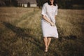 Stylish girl in linen dress and hat running barefoot in grass in sunny field at village. Boho woman relaxing in countryside, Royalty Free Stock Photo