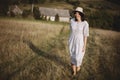 Stylish girl in linen dress and hat walking barefoot in grass in sunny field at village. Boho woman relaxing in countryside, Royalty Free Stock Photo