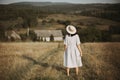 Stylish girl in linen dress and hat walking barefoot in grass in sunny field at village. Boho woman relaxing in countryside, Royalty Free Stock Photo
