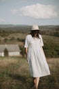 Stylish girl in linen dress and hat walking barefoot among herbs and wildflowers in sunny field in mountains. Boho woman relaxing Royalty Free Stock Photo