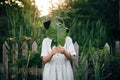 Stylish girl in linen dress holding big green leaf at face at wooden fence and grass. Portrait of boho woman in hat posing with Royalty Free Stock Photo