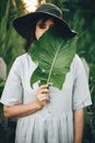 Stylish girl in linen dress holding big green leaf at face at wooden fence and grass. Portrait of boho woman in hat posing with Royalty Free Stock Photo