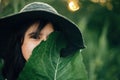 Stylish girl in linen dress holding big green leaf at face at wooden fence and grass. Portrait of boho woman in hat posing with Royalty Free Stock Photo