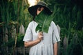 Stylish girl in linen dress holding big green leaf at face at wooden fence and grass. Portrait of boho woman in hat posing with Royalty Free Stock Photo