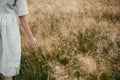 Stylish girl in linen dress holding hand among herbs and wildflowers in field. Boho woman walking in countryside among grass,