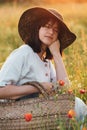 Stylish girl in linen dress sitting in poppy meadow in sunset with flowers in rustic straw basket. Boho woman  in hat relaxing in Royalty Free Stock Photo