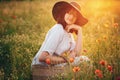 Stylish girl in linen dress sitting in poppy meadow in sunset with flowers in rustic straw basket. Boho woman  in hat relaxing in Royalty Free Stock Photo