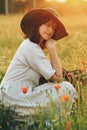 Stylish girl in linen dress sitting in poppy meadow in sunset with flowers in rustic straw basket. Boho woman  in hat relaxing in Royalty Free Stock Photo