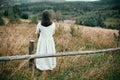 Stylish girl in linen dress standing at aged wooden fence among herbs and wildflowers, looking at field. Boho woman relaxing in