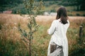 Stylish girl in linen dress standing at aged wooden fence among herbs and wildflowers, looking at field. Boho woman relaxing in