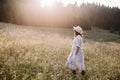 Stylish girl in linen dress walking among wildflowers in sunny meadow in mountains. Boho woman relaxing in countryside flowers at Royalty Free Stock Photo