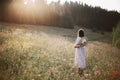 Stylish girl in linen dress walking among wildflowers in sunny meadow in mountains. Boho woman relaxing in countryside flowers at Royalty Free Stock Photo