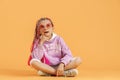 Stylish girl in rounded glasses with pink dreadlocks sitting, thinking, look up and posing on a yellow background Royalty Free Stock Photo