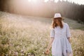 Stylish girl in rustic dress and hat walking among wildflowers in sunny meadow in mountains. Boho woman relaxing in countryside Royalty Free Stock Photo