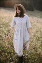 Stylish girl in rustic dress and hat walking among wildflowers in sunny meadow in mountains. Boho woman relaxing in countryside Royalty Free Stock Photo