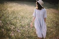 Stylish girl in rustic dress and hat walking among wildflowers in sunny meadow in mountains. Boho woman relaxing in countryside Royalty Free Stock Photo