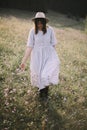 Stylish girl in rustic dress walking in wildflowers in sunny meadow in mountains. Boho woman relaxing in countryside flowers at Royalty Free Stock Photo