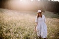 Stylish girl in rustic dress walking in wildflowers in sunny meadow in mountains. Boho woman relaxing in countryside flowers at Royalty Free Stock Photo