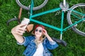 Stylish girl in the sunglasses and a denim jacket lays on a lawn next to a bike and takes selfie on a smartphone Royalty Free Stock Photo
