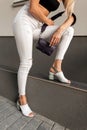 Stylish girl in trendy white jeans in black summer shoes with bag posing near gray  wall on the street. Close-up Royalty Free Stock Photo