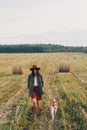 Stylish girl walking with her dog at hay bale in summer field in sunset. Young woman in hat traveling with pet in haystack field.